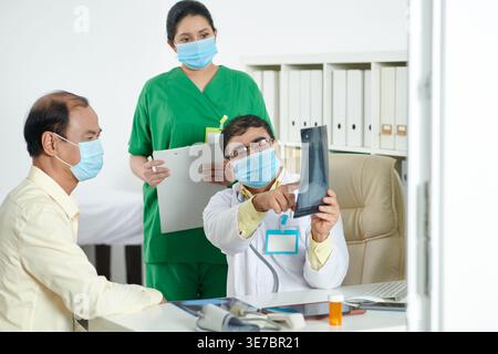 Indischer männlicher Doktor mit asiatischer Krankenschwester neben ihm, die Gesichtsmasken trägt, die dem asiatischen Mann Röntgenergebnisse zeigen, während er am Tisch sitzt... Stockfoto