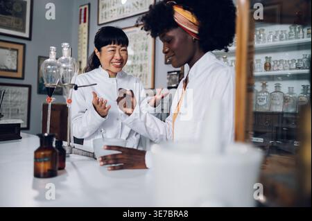Japanische Chemikerin und schwarze Kollegin, die an Experimenten in der Vintage-Apotheke steht und arbeitet Stockfoto