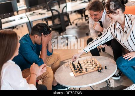 Enttäuschter Mann aus dem Nahen Osten, der das Spiel verlor, das sein Gesicht mit Händen bedeckt und Schach spielt, mit kaukasischen Arbeitskollegen, die im Büro auf dem Stuhl sitzen... Stockfoto