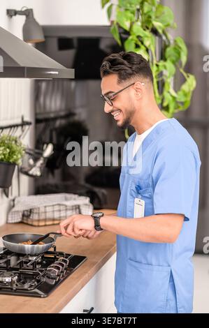 Lächelnder arabischer männlicher Pfleger in Brille kocht, während er zu Hause in der Küche steht Stockfoto