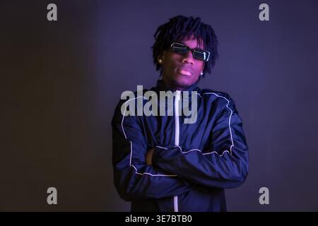 Ernster Schwarzer Mann mit Wendungen, der schwarze Jacke und Sonnenbrille trägt und im Studio mit überkreuzten Armen vor dunklem Hintergrund steht Stockfoto