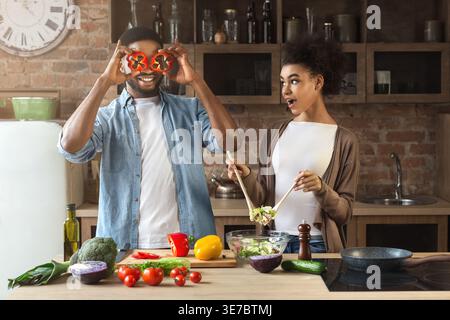 Lächelndes schwarzes Paar, das in Scheiben geschnittene Paprika hält und Salat zubereitete, während er am Tisch in der Küche zu Hause steht Stockfoto