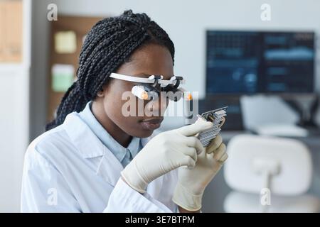 Fokussierte schwarze Computeringenieurin mit Zöpfen, die eine Lupe tragen und elektronische Teile untersuchen, die im Techniklabor am Tisch sitzen Stockfoto