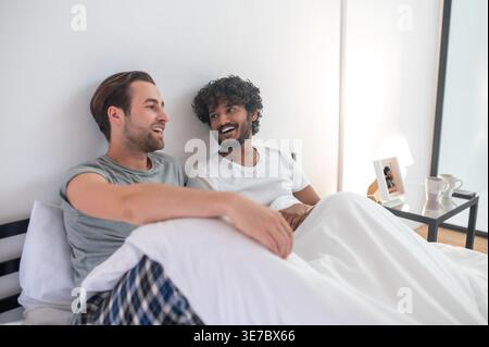 Lächelndes indisches und kaukasisches schwules Paar, das im Bett sitzt Stockfoto
