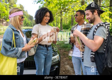 Eine lächelnde Gruppe verschiedener Freunde isst Sandwiches, während sie tagsüber zusammen auf der Waldstraße stehen Stockfoto