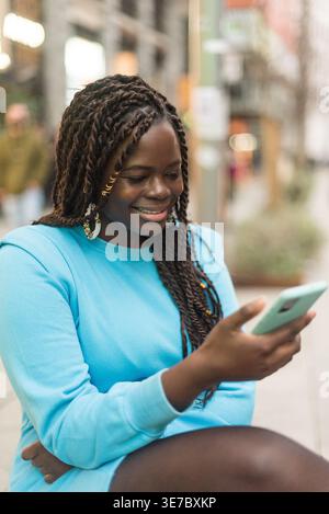 Lächelnde schwarze Frau mit Wendungen, die einen blauen Pullover mit Smartphone trägt, während sie auf der Stadtstraße steht Stockfoto