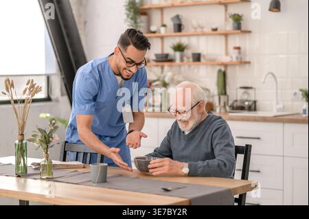Lächelnder arabischer männlicher Pfleger mit Brille, der Essen in der Nähe eines älteren Mannes gibt, während er zu Hause am Tisch sitzt Stockfoto