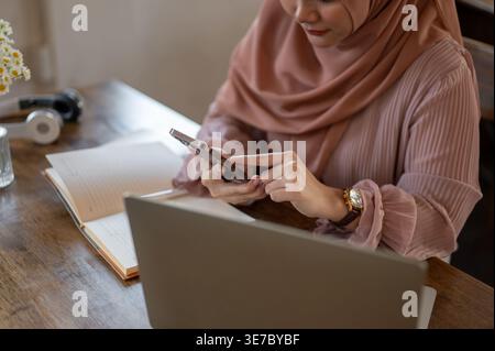 Asiatische muslimische Frau im Hijab mit Smartphone am Tisch mit Laptop und Notebook im Café Stockfoto
