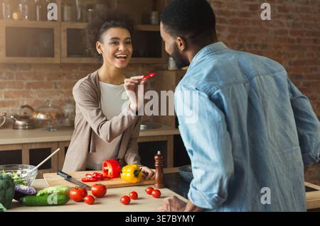Lächelnde Schwarze Freundin füttert Schwarzen Freund, während sie am Tisch mit Gemüse in der Küche zu Hause steht Stockfoto