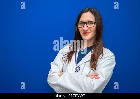 Professionelle Ärztin mit weißem Mantel und Stethoskop, selbstbewusst lächelnd vor blauem Hintergrund, repräsentiert das Gesundheitswesen Stockfoto
