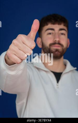 Ein lächelnder bärtiger Mann gibt die Daumen hoch für Anerkennung und Erfolg, Porträt auf blauem Hintergrund vermittelt Selbstvertrauen und positive Emotionen Stockfoto