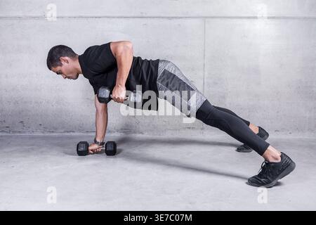 Hispanischer Mann mit Kurzhanteln, die aus Liegestützen auf grauem Beton rudern Stockfoto