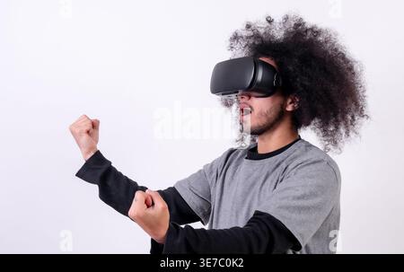 Hispanischer Mann mit lockigem Haar, der im Studio eine VR-Brille mit geballten Fäusten vor weißem Hintergrund trägt Stockfoto