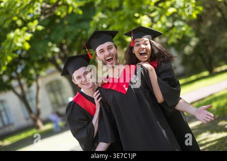Fröhliche schwarze Frau in akademischem Kleid und Mütze, die zusammen mit kaukasischen männlichen Freunden steht und Spaß auf dem Schulgelände am Abschlussfeiertag hat Stockfoto