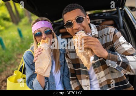 Schwarzes und kaukasisches Paar mit Sonnenbrille, das tagsüber Sandwiches im offenen Kofferraum auf der Forststraße isst Stockfoto