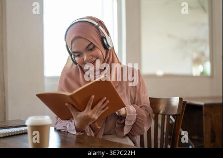 Lächelnde asiatische muslimische Frau, die Hijab trägt und Musik mit Kopfhörern hört, während sie im Café am Tisch sitzt Stockfoto