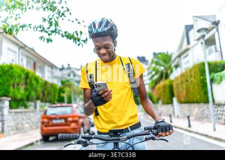 Schwarzer Mann mit Helm und Liefertasche, der den Kunden mit dem Smartphone kontaktiert, während er tagsüber mit dem Fahrrad auf der Straße steht Stockfoto