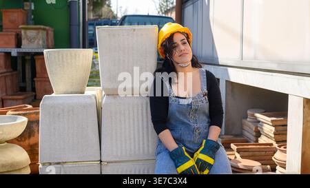 Ernsthafte hispanische Arbeiterin mit Denim-Overall und gelbem Schutzhelm, die sich während des brückens auf weißen Töpfen auf der Baustelle zurücklehnen... Stockfoto