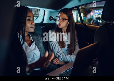 Ernste Schwarze Frau mit Flechten, die einen Laptop benutzt und sich mit einer kaukasischen Kollegin trifft, die während der Reise auf dem Rücksitz des Autos sitzt Stockfoto