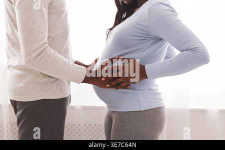Zugeschnittenes Bild eines Schwarzen Mannes, der den Bauch einer schwangeren Frau berührt, die sich tagsüber im Schlafzimmer gegenübersieht Stockfoto