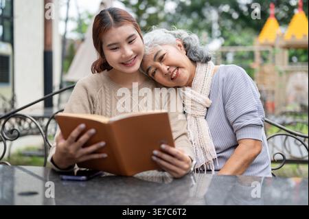 Thai Senior Frau, die Spaß beim Lesen von Büchern mit ihrer Enkelin hat, während sie tagsüber am Tisch im Garten sitzt Stockfoto
