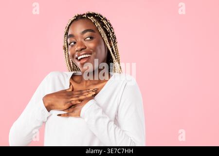 Schwarze Frau, die Hände auf der Brust hält und drückt, die seitlich vor rosa Hintergrund im Studio steht Stockfoto