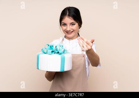 Hispanische Konditorin, die im Studio eine einladende Handgeste mit Kuchen vor beigefarbenem Hintergrund hält Stockfoto