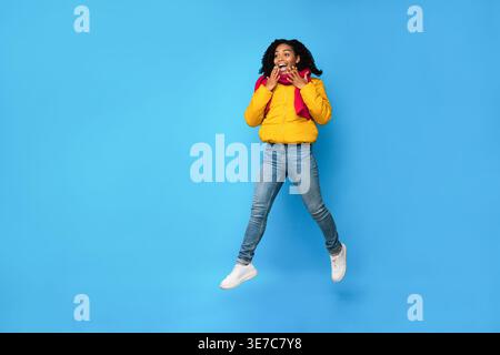 Überraschte schwarze Frau in Winterjacke und Schal, die im Studio vor blauem Hintergrund hüpft Stockfoto