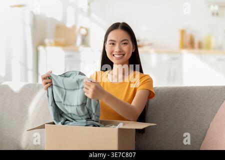 Asiatische Frau, die Parce neues Hemd aus der Box auspackt, sitzend auf Sofa im Wohnzimmer zu Hause Stockfoto