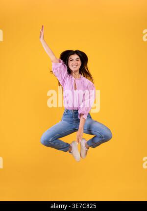 Lächelnde armenische Frau springt hoch und berührt die Beine mit der Hand vor gelbem Hintergrund im Studio Stockfoto