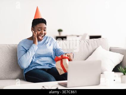 Überraschte schwarze Frau mit kurzem Afro tragendem Partyhut, der eine Geschenkbox hält, während sie Videoanrufe über Laptop im Wohnzimmer zu Hause hatte Stockfoto