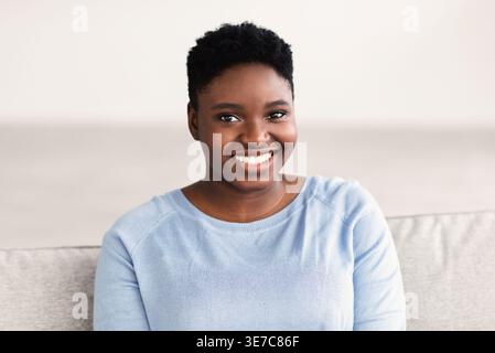Lächelnde schwarze Frau mit kurzem Afro, die zu Hause im Wohnzimmer auf der Couch sitzt Stockfoto