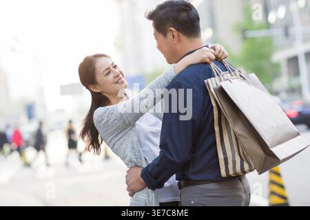Fröhliches chinesisches Paar mittleren Alters, das sich umarmt und ansieht, während es tagsüber mit Einkaufstaschen auf der Stadtstraße steht Stockfoto