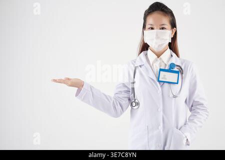 Asiatische Ärztin mit chirurgischer Maske Stethoskop um den Hals Hand in Tasche und etwas in der Hand halten, während sie vor weißem Hintergrund steht... Stockfoto