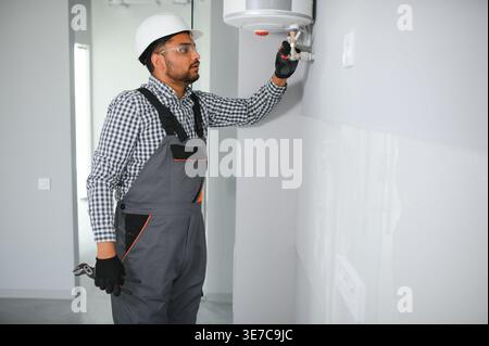 Indischer Klempner mit Uniform und Schutzhelm, der im Bad an der Heizung steht und arbeitet Stockfoto
