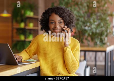 Schwarze Frau mit lockigen Haaren, die telefoniert, am Tisch mit Laptop im Café Stockfoto