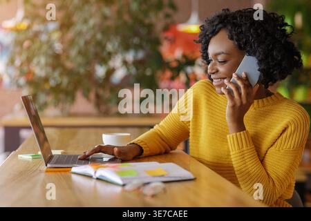 Schwarze Frau mit lockigen Haaren, die telefoniert, während sie mit einem Laptop am Tisch sitzt, mit Notebook und Kaffeetasse im Café Stockfoto