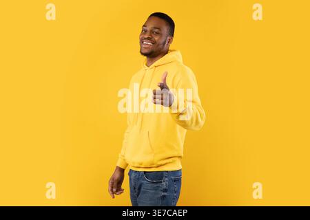 Lächelnder bärtiger Schwarzer Mann mit Hoodie, der den Finger nach vorne zeigt, während er seitlich vor gelbem Hintergrund im Studio steht Stockfoto