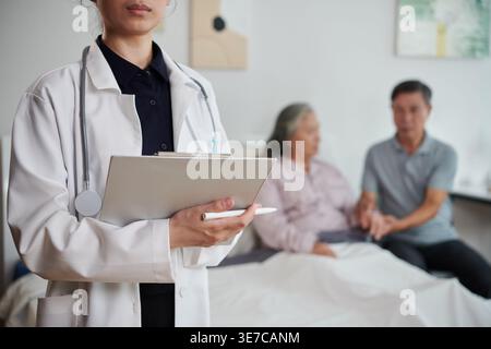 Zugeschnittenes Bild des asiatischen weiblichen Arztes, der die medizinischen Aufzeichnungen des Patienten füllt, nachdem sie eine ältere Frau mit ihrem Mann überprüft hat, der auf dem Bett von Hos sitzt... Stockfoto