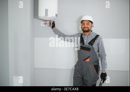 Indischer Klempner mit Uniform und Schutzhelm, der im Bad an der Heizung steht und arbeitet Stockfoto