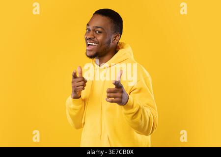 Lächelnder bärtiger Schwarzer Mann mit Hoodie, der die Finger nach vorne zeigt, während er seitlich vor gelbem Hintergrund im Studio steht Stockfoto