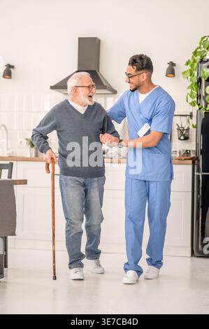 Lächelnder arabischer männlicher Betreuer in Brille, der einen älteren Mann im Stehen zu Hause ansieht Stockfoto
