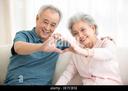 Lächelndes chinesisches Seniorenpaar, das Herz Hand Zeichen macht, während es auf Sofa zu Hause im Wohnzimmer sitzt Stockfoto