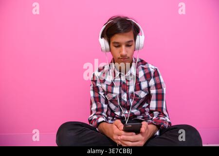 Ernster männlicher Teenager aus dem Nahen Osten in karierten langen Ärmeln, der Musik über Kopfhörer mit Smartphone hört, während er auf dem Boden gegen die Nadel sitzt... Stockfoto