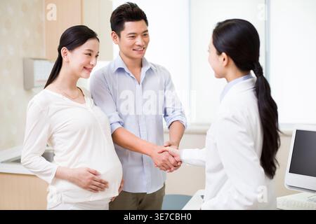 Lächelnder chinesischer Mann schüttelt der chinesischen Ärztin die Hände, während er mit einer chinesischen schwangeren Frau im Krankenhaus steht Stockfoto