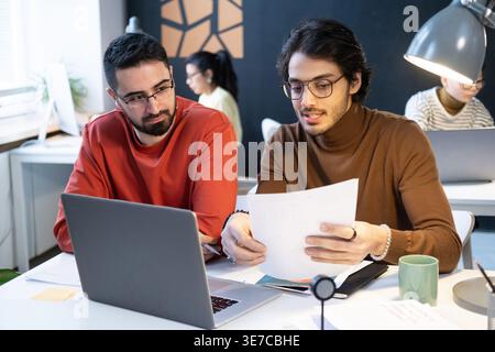 Geschäftsleute aus dem Nahen Osten diskutieren und halten Dokumente in der Hand, während sie am Tisch mit Laptop in der Nähe von Kollegen im Büro sitzen Stockfoto