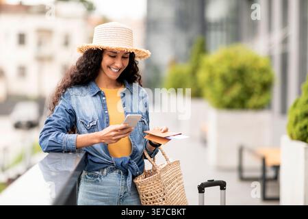 Frau aus dem Nahen Osten mit Strohhut mit Smartphone und Flugunterlagen, die am Flughafen mit Rattanhandtasche und Koffer stehen Stockfoto
