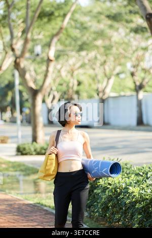 Vietnamesische Frau trägt Sonnenbrille und Fitness Tragetasche mit Yogamatte, die am sonnigen Tag im Park spazieren geht Stockfoto
