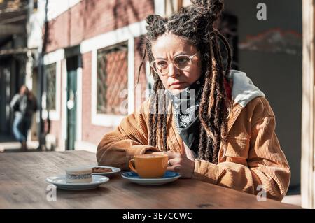 Hispanische Frau mit Dreadlocks, die eine Brille trug und wegblickte, während sie tagsüber im Café im Freien mit einer Tasse Kaffee und Gebäck am Tisch saß Stockfoto