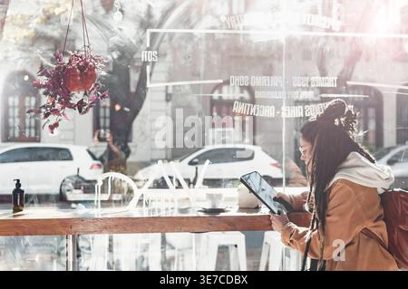 Hispanische Frau mit Dreadlocks, die eine Brille trägt, die ein Tablet hält, während sie tagsüber am Tisch im Café im Freien sitzt Stockfoto